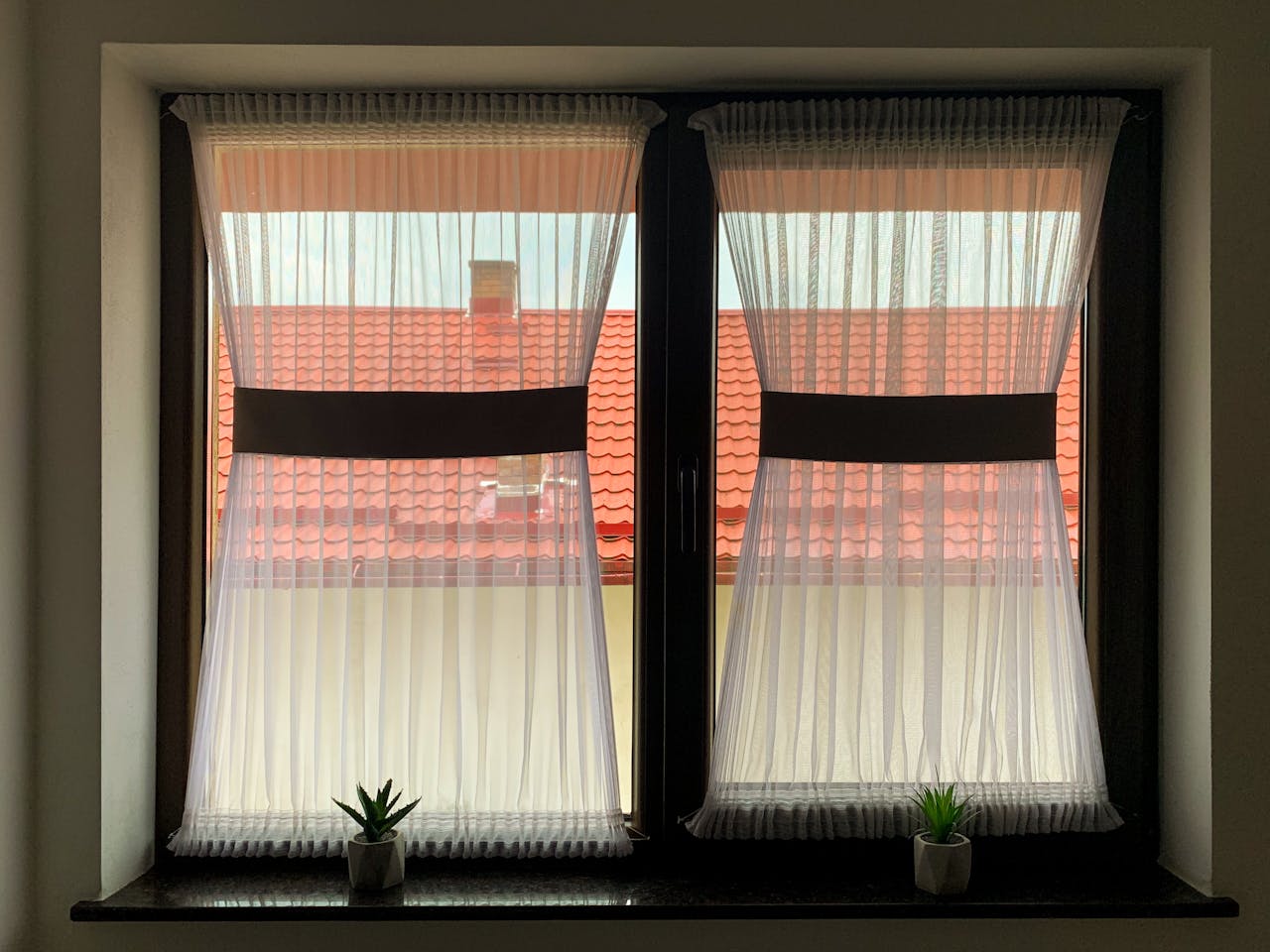 A window with sheer white curtains, red roof view, and small potted plants on the sill.